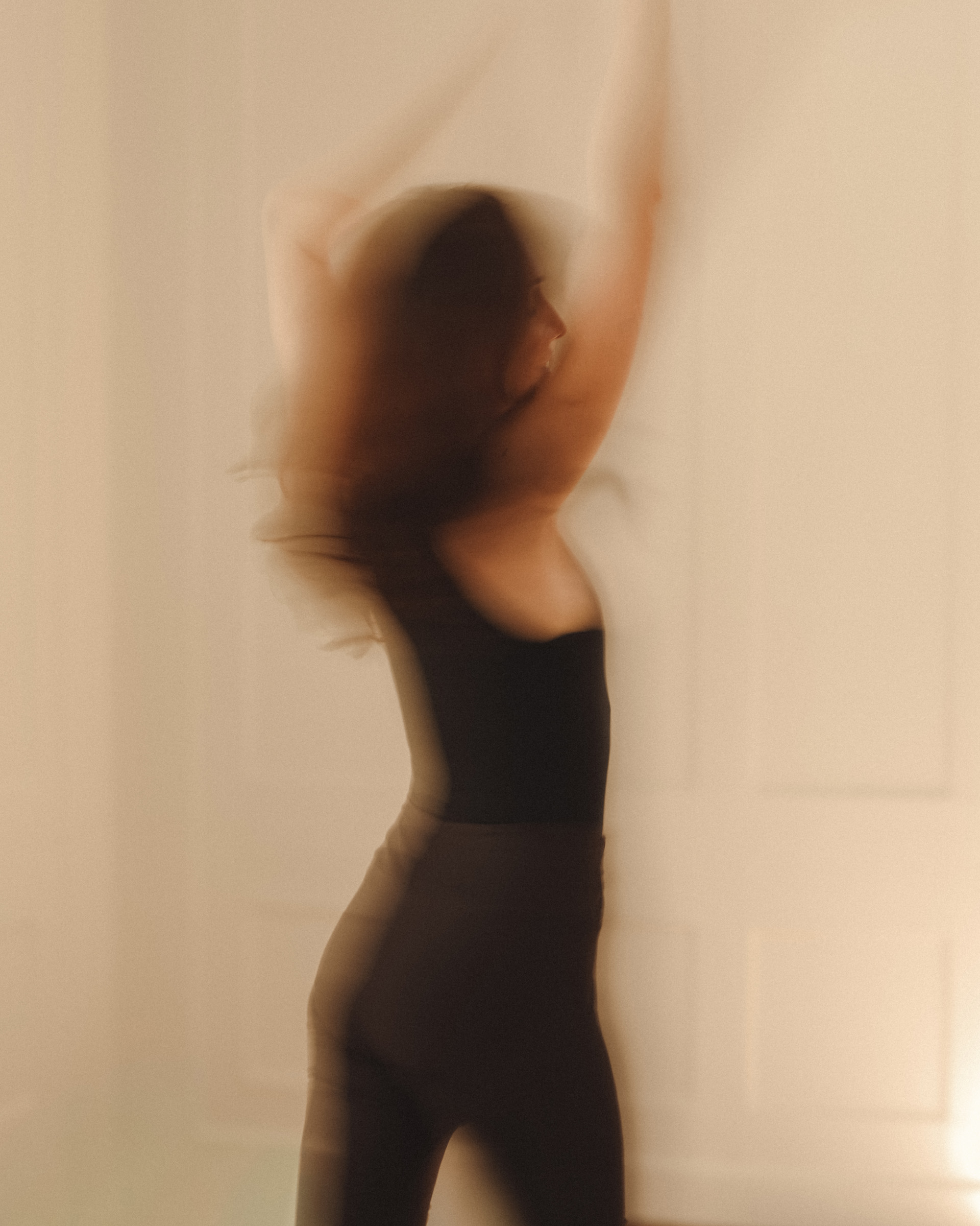 Femme en débardeur et legging noirs les bras levés en mouvement, silhouette floue sur fond clair évoquant la danse ou le yoga.