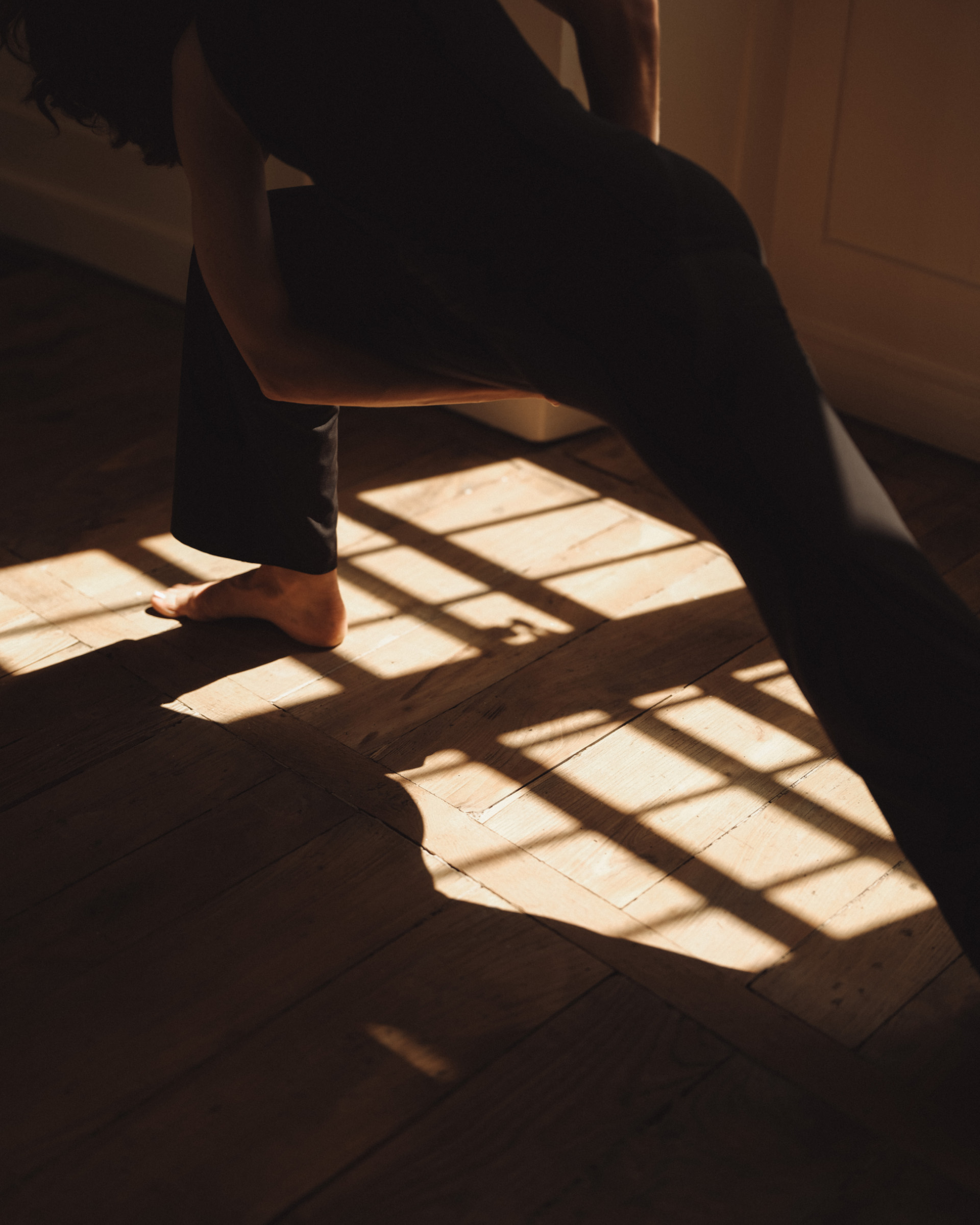Femme en débardeur et legging évasé noirs en mouvement dans une position de yoga, pieds nus sur un parquet en bois ancien avec jeu d'ombres géométriques et lumière dorée filtrée par une fenêtre à croisillons.